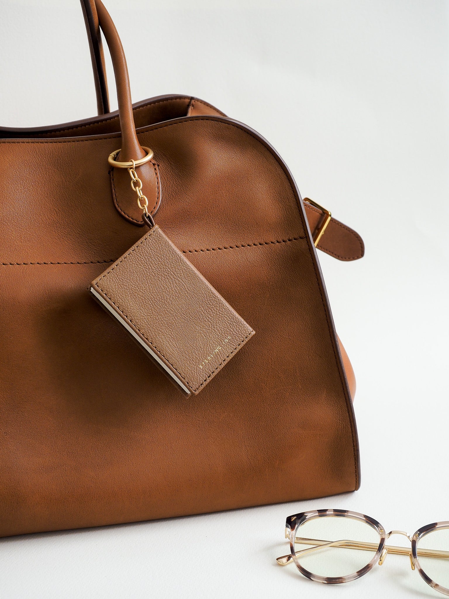 A close-up of a brown leather handbag with a matching leather luggage tag attached by gold hardware, photographed on a clean white background. A pair of tortoiseshell eyeglasses is partially visible in the lower right corner.
#Color_Caramel