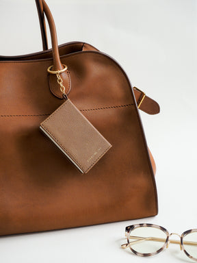 A close-up of a brown leather handbag with a matching leather luggage tag attached by gold hardware, photographed on a clean white background. A pair of tortoiseshell eyeglasses is partially visible in the lower right corner.
#Color_Caramel