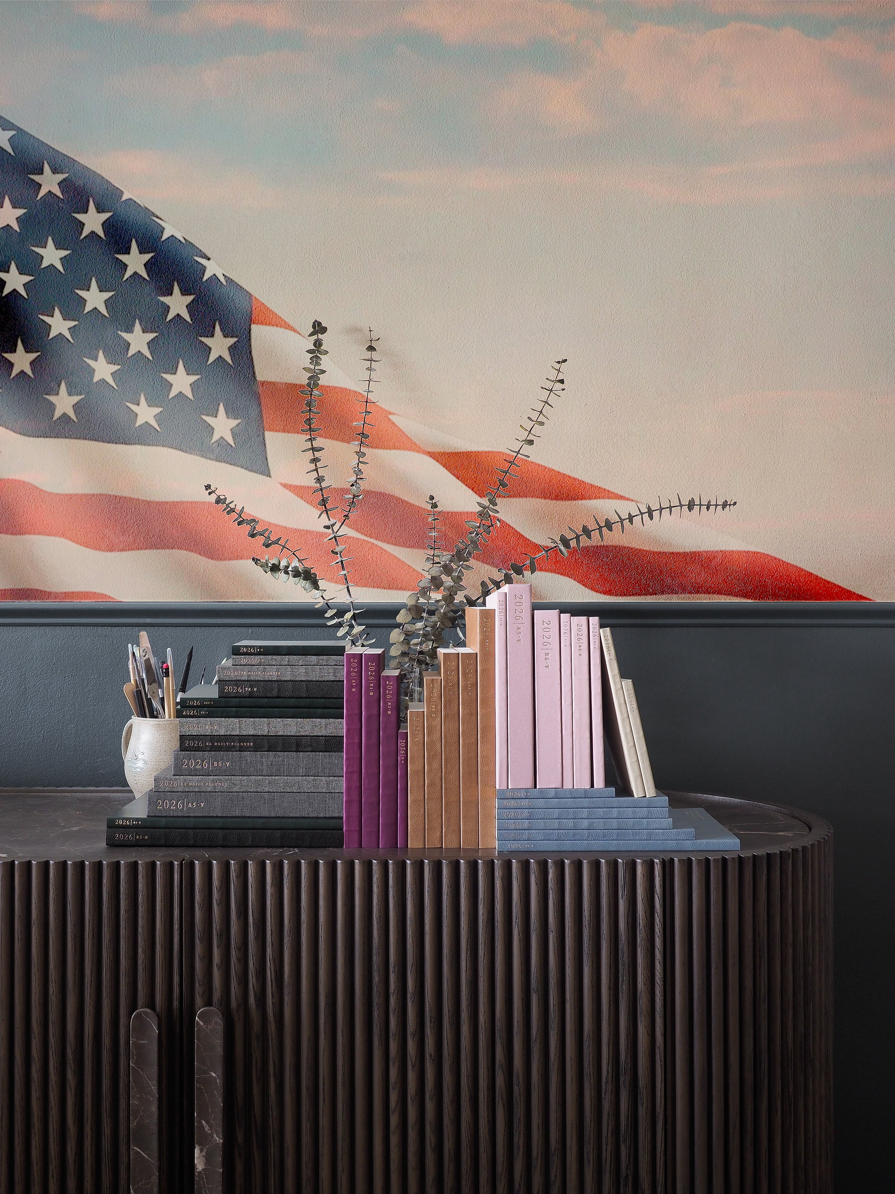 Stacked pastel-colored planners, journals, and assorted writing supplies are displayed on a dark dresser against a background of a stylized American flag.
