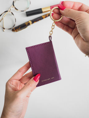 A person holds a small purple leather card holder keychain with a gold split ring and chain. The card holder has gold stitching and the words 'STELLING INK' printed in gold near the bottom.
#Color_Mulberry