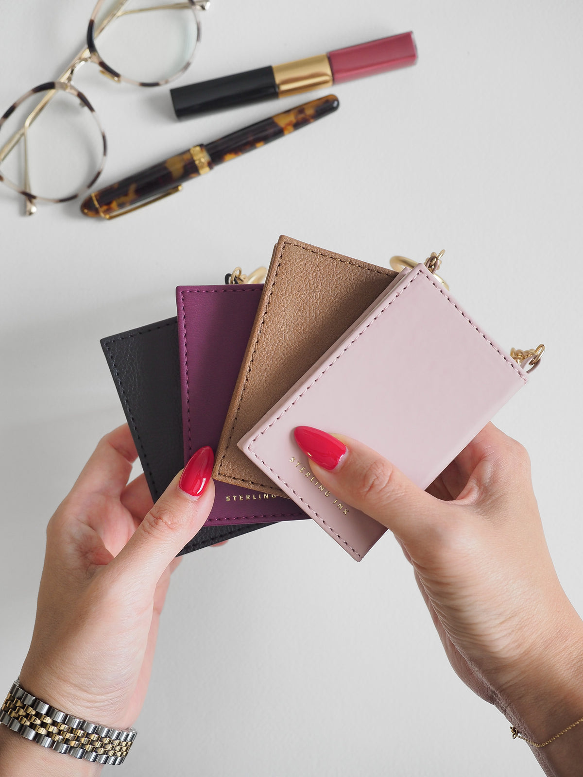 Four small leather card holders or wallet-style pouches in black, purple, tan, and blush pink are fanned out in a hand-held product shot. Two hands with bright pink nail polish hold the wallets, with gold-tone jewelry and cosmetics visible on a light gray surface.