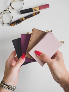 Four small leather card holders or wallet-style pouches in black, purple, tan, and blush pink are fanned out in a hand-held product shot. Two hands with bright pink nail polish hold the wallets, with gold-tone jewelry and cosmetics visible on a light gray surface.