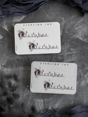 Cream-colored "October" month sticker featuring a rustic, distressed vintage design with dark ink lettering and small decorative bat silhouettes perfect for a fall theme.