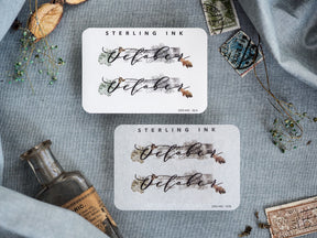 Elegant "October" month stickers with flowing script typography accented by rustic, muted watercolor washes and autumnal floral sprigs.