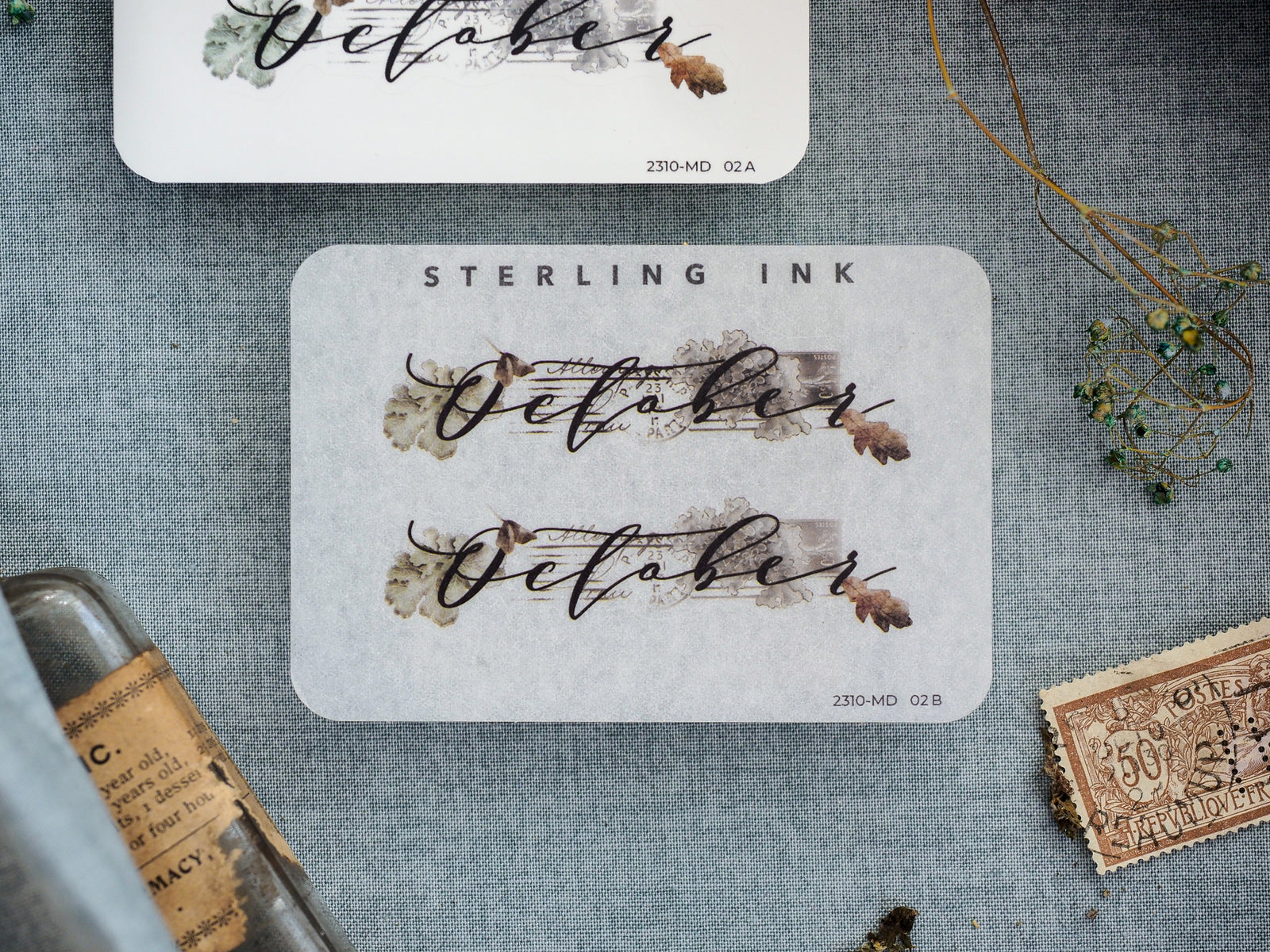 Rustic "October" month sticker featuring elegant script typography and delicate sepia-toned botanical accents on a clean white background.