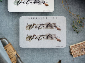 Rustic "October" month sticker featuring elegant script typography and delicate sepia-toned botanical accents on a clean white background.