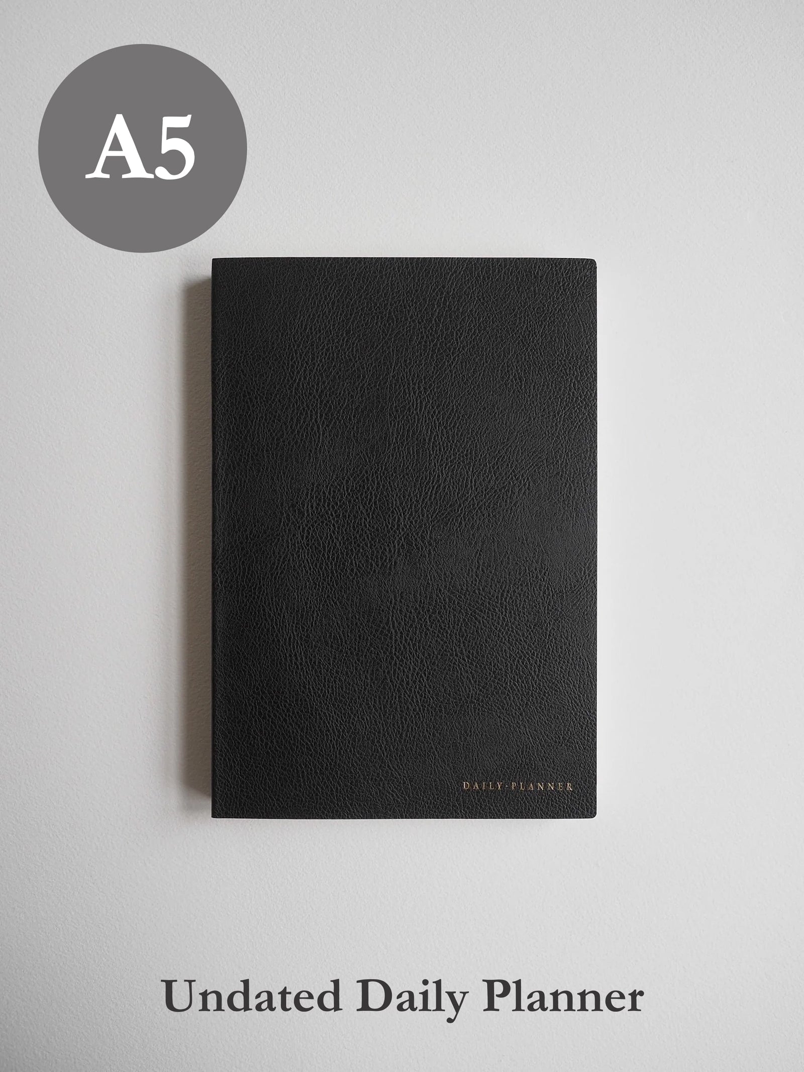 Sleek A5 undated daily planner bound in matte black material, featuring discreet gold foiled lettering near the bottom center.