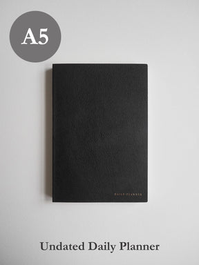 Sleek A5 undated daily planner bound in matte black material, featuring discreet gold foiled lettering near the bottom center.