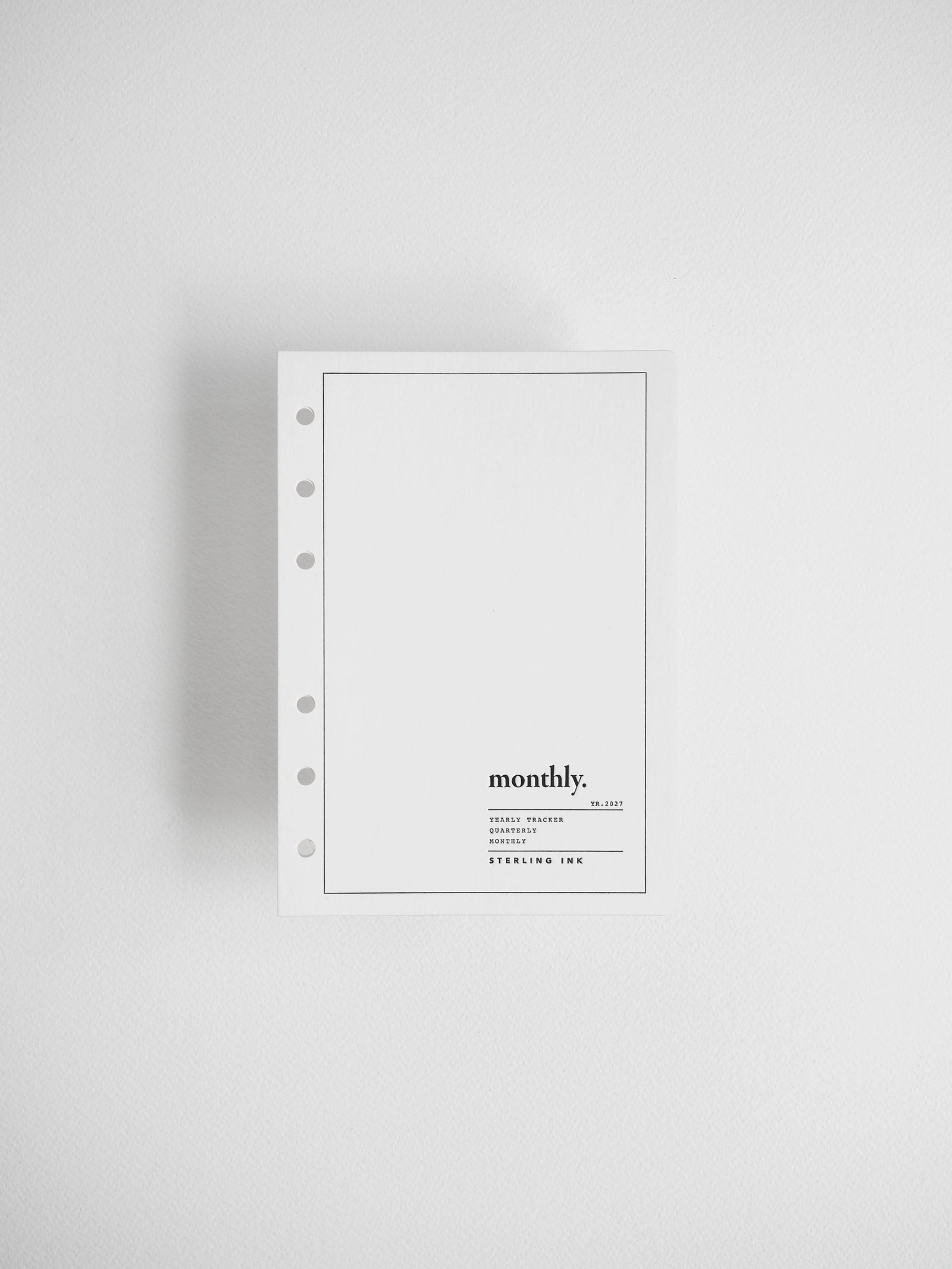 A minimalist white monthly binder notebook with five-hole punch on the left side, featuring a thin black border and the text 'monthly.' with smaller text below, displayed on a clean light gray background.
#Size_A6