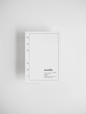 A minimalist white monthly binder notebook with five-hole punch on the left side, featuring a thin black border and the text 'monthly.' with smaller text below, displayed on a clean light gray background.
#Size_A6