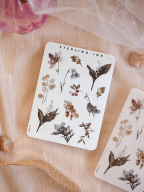 Elegant sticker sheet featuring muted botanical elements, including delicate dried foliage, small berries, and stylized flowers in sepia, dusty brown, and deep indigo tones.