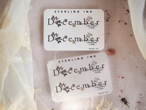 Set of white calendar stickers celebrating December, featuring black cursive script and delicate pressed floral accents perfect for journaling and planners.