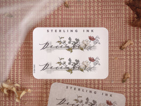 Elegant December month sticker featuring the word in scripted lettering and decorated with delicate watercolor illustrations of blooming flowers and green foliage.