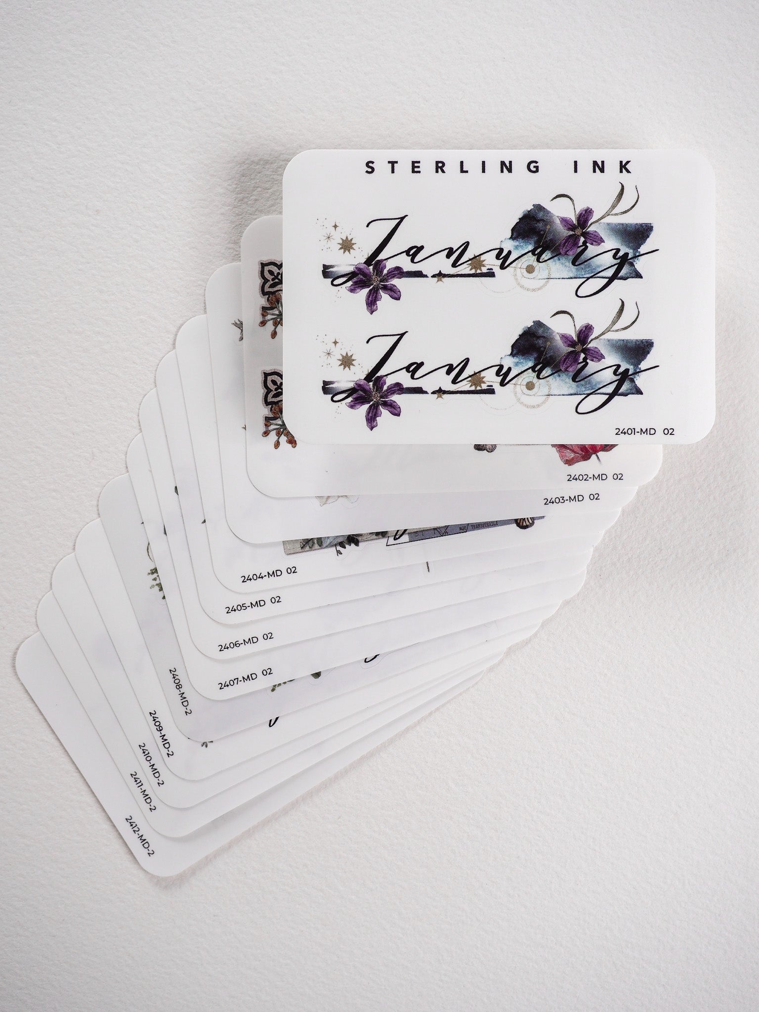 Decorative monthly sticker set featuring elegant floral illustrations, deep purple and blue watercolor blooms, and graceful script calligraphy for each month.