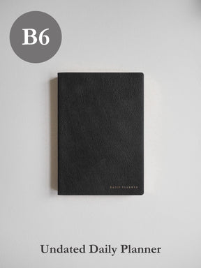 Sleek B6 undated daily planner featuring a textured, dark charcoal matte cover with minimal, elegant branding detail.