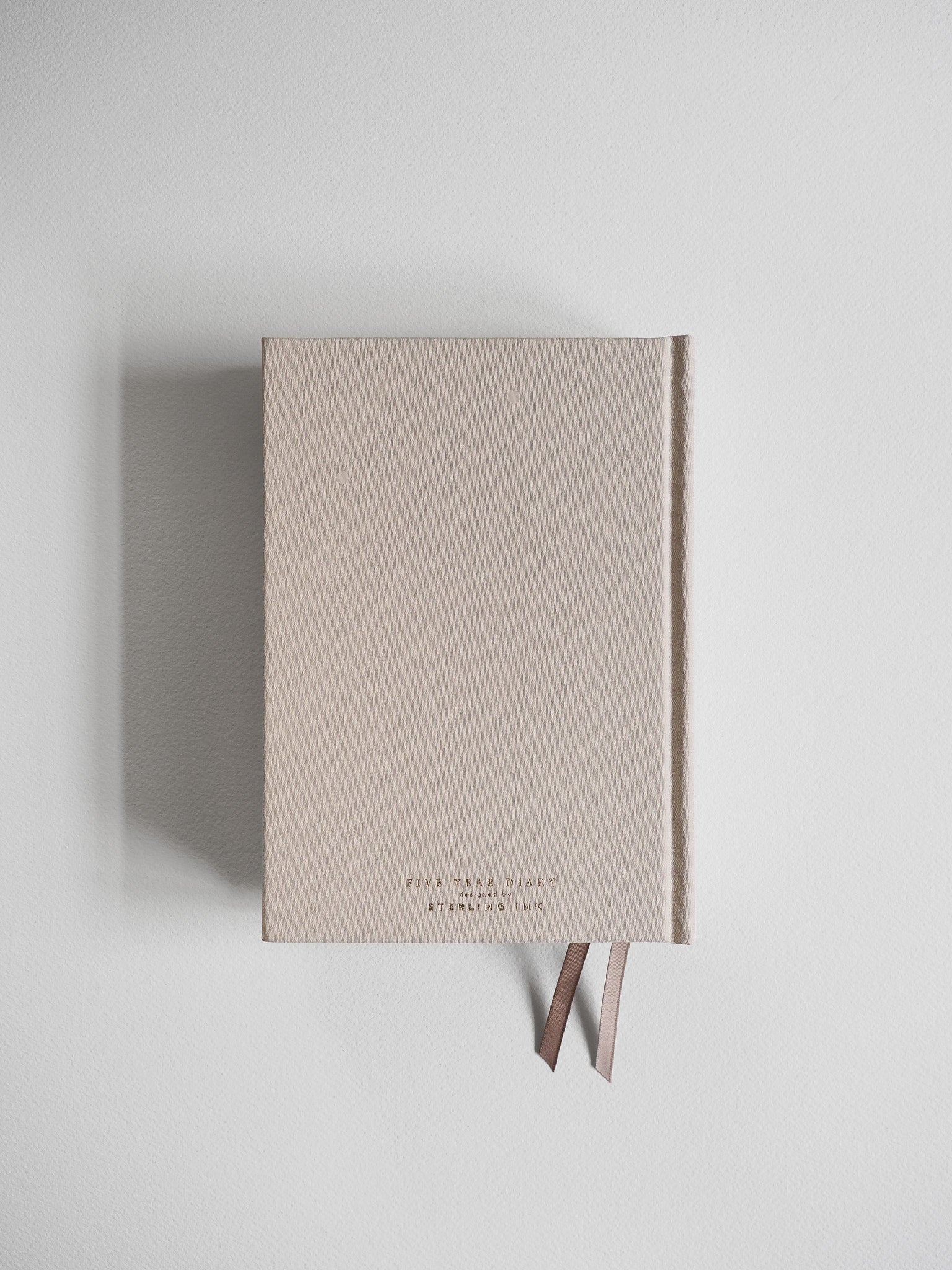 Minimalist five-year diary with a neutral light beige hardcover, featuring debossed branding and held closed with an elegant brown fabric ribbon tie.