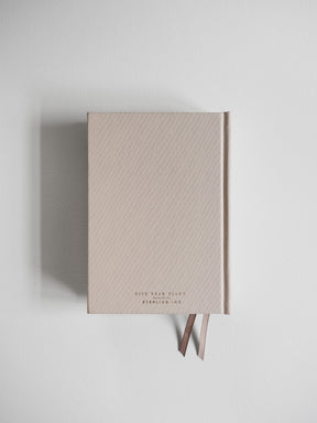 Minimalist five-year diary with a neutral light beige hardcover, featuring debossed branding and held closed with an elegant brown fabric ribbon tie.
