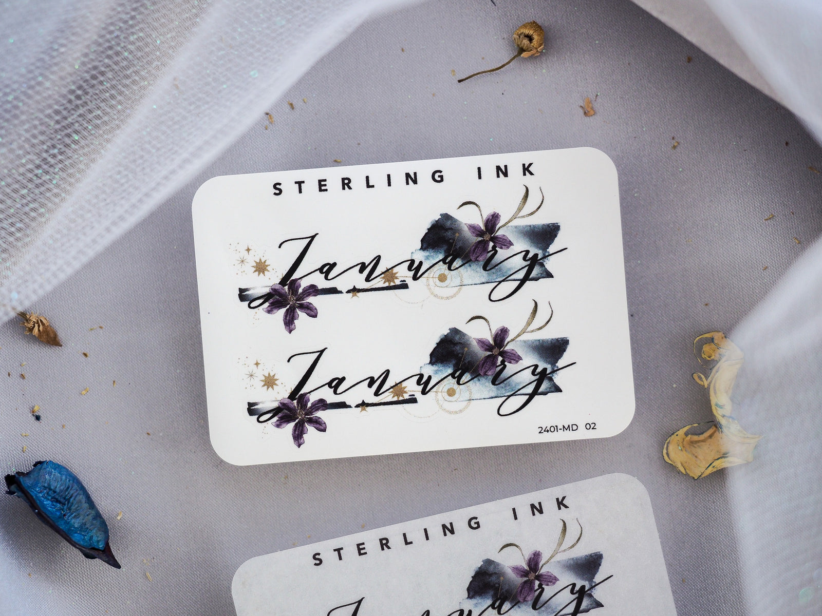 White sticker featuring the scripted word "January" and "Sterling Ink," adorned with delicate dark purple and navy floral illustrations and gold accents.