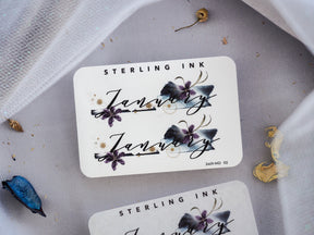 White sticker featuring the scripted word "January" and "Sterling Ink," adorned with delicate dark purple and navy floral illustrations and gold accents.