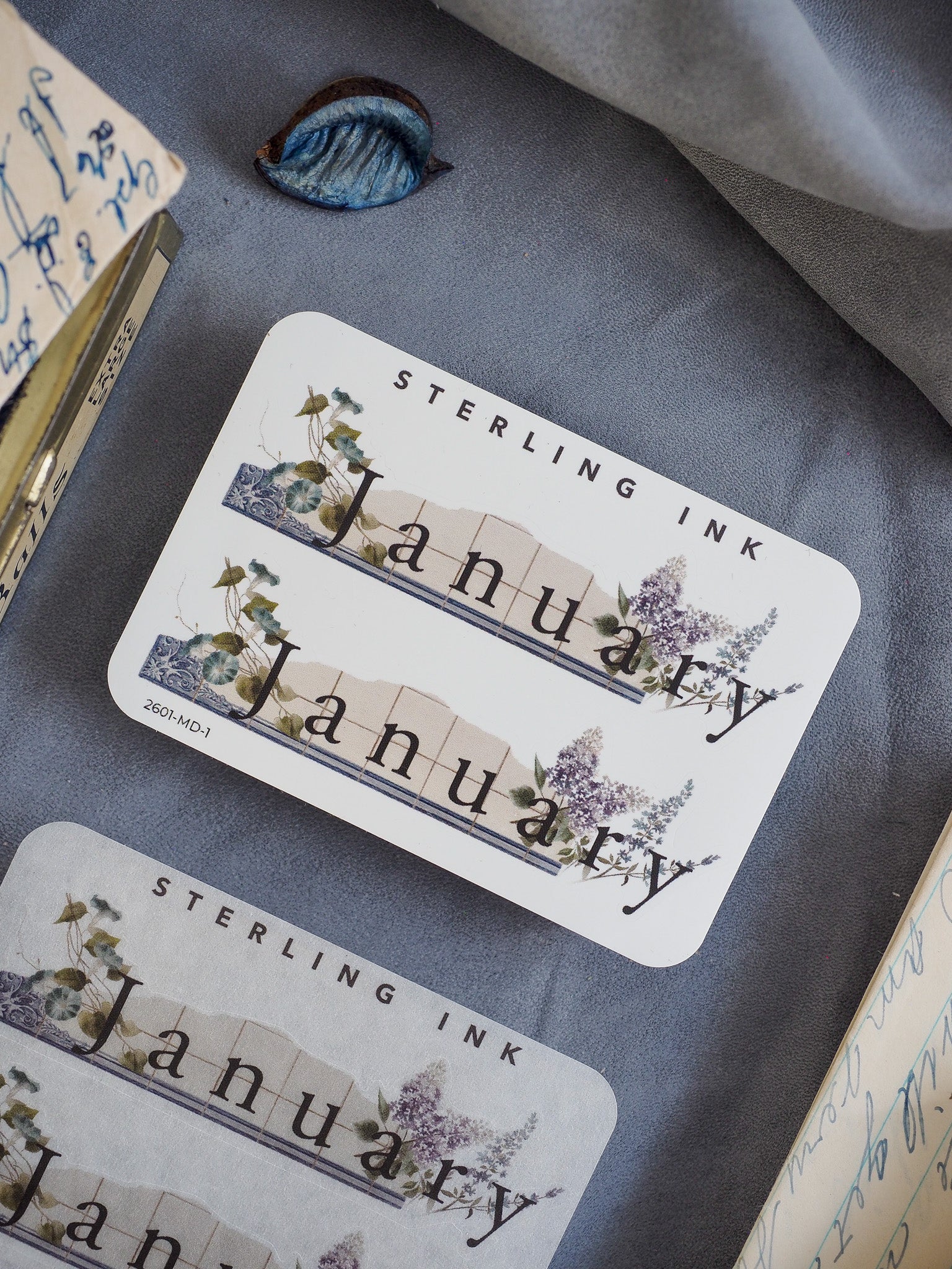 Watercolor-themed January month sticker featuring "Sterling Ink" typography, framed by delicate watercolor illustrations of lush green foliage and soft purple blooms.