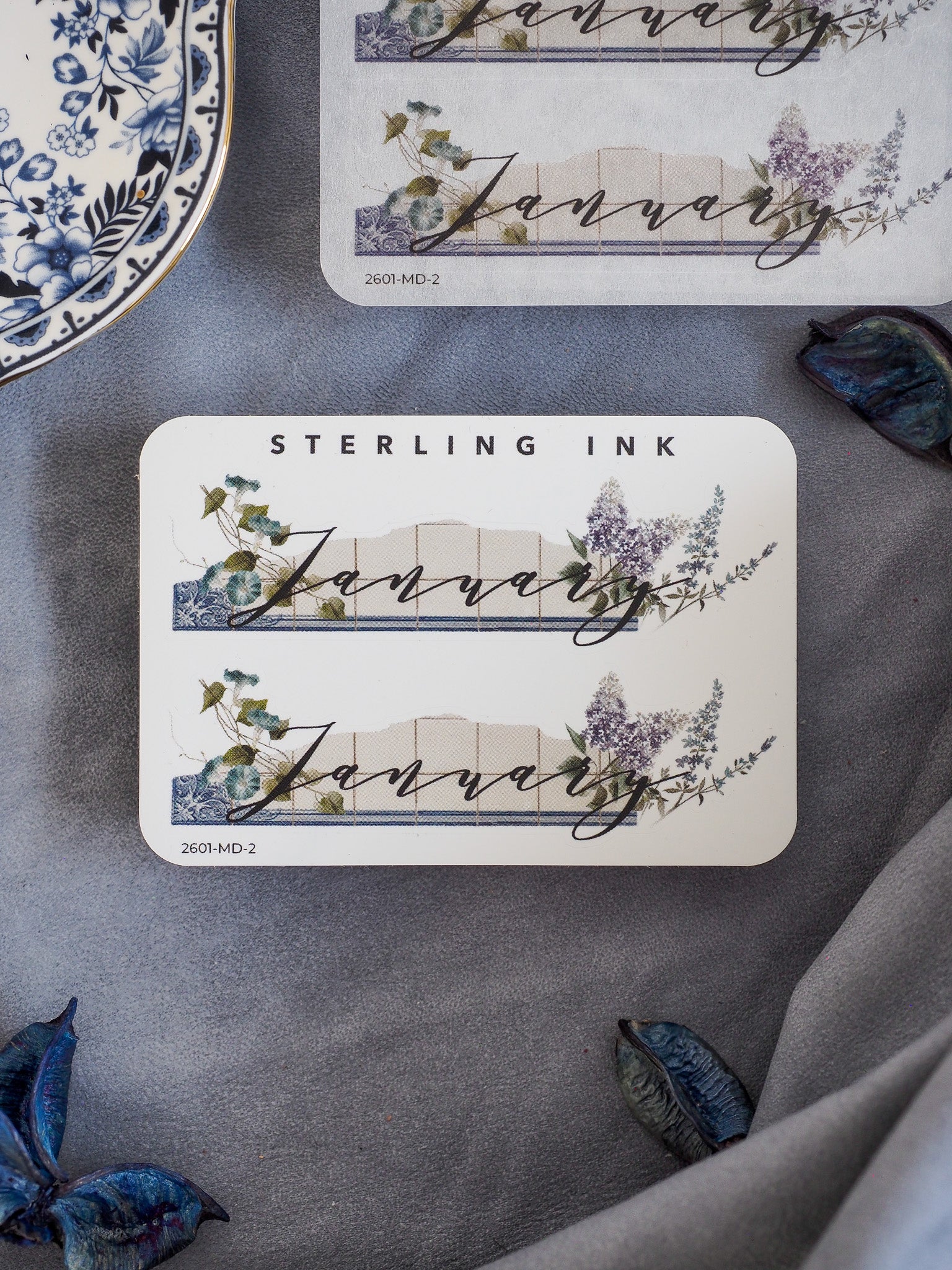 Watercolor month sticker for January featuring the word "JANUARY" centered over a banner and framed by delicate green and purple floral wreaths.