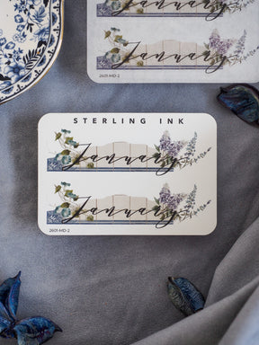 Watercolor month sticker for January featuring the word "JANUARY" centered over a banner and framed by delicate green and purple floral wreaths.