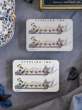 Elegant "January" month sticker featuring scripted text over a delicate watercolor illustration of blue foliage and purple wildflowers.