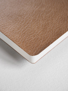 Close-up of a notebook corner showing a tan textured cover and a white page edge on a light gray background.
#Color_Caramel