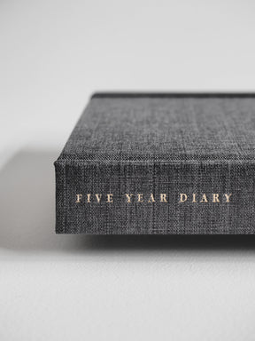 Dark charcoal hardcover diary featuring a woven linen texture and the title "FIVE YEAR DIARY" discreetly printed in small, elegant metallic letters.