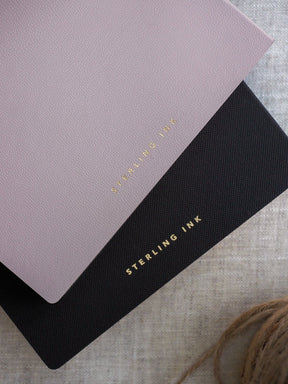 Two stacked grid notebooks, one blush pink and one black, featuring gold foiled "STEARLING INK" branding for a cohesive, elegant design.