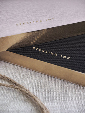 Open notebook displaying an off-white cover with elegant gold foiled branding and a contrasting dark page featuring a neat grid pattern for writing.