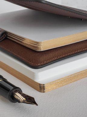Stack of pristine, gold-edged lined loose leaf paper, presented with dark brown leather-look accents, ideal for journaling or record-keeping.