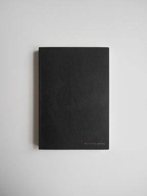 Matte black A5 daily planner with clean lines and a refined, minimal aesthetic, featuring delicate gold foil lettering on the solid cover.