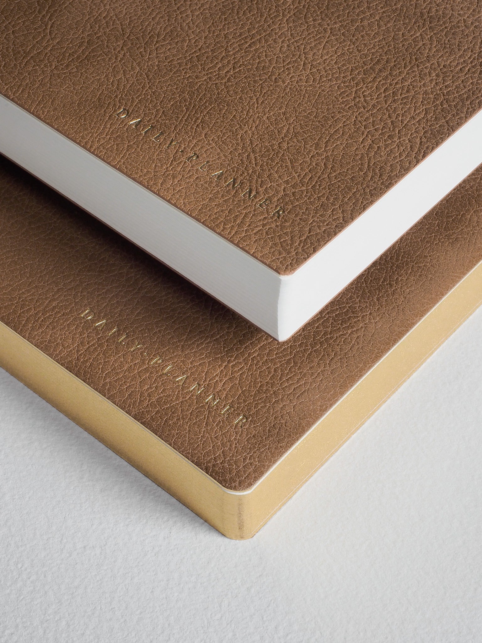A stack of A5 daily planners featuring rich tan and taupe textured faux-leather covers, showcasing elegant embossed branding and crisp white edges.