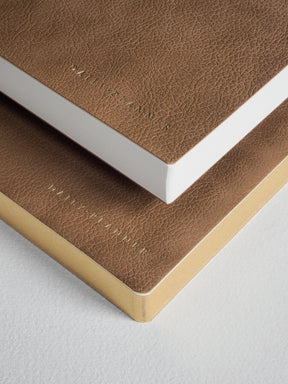 A stack of A5 daily planners featuring rich tan and taupe textured faux-leather covers, showcasing elegant embossed branding and crisp white edges.