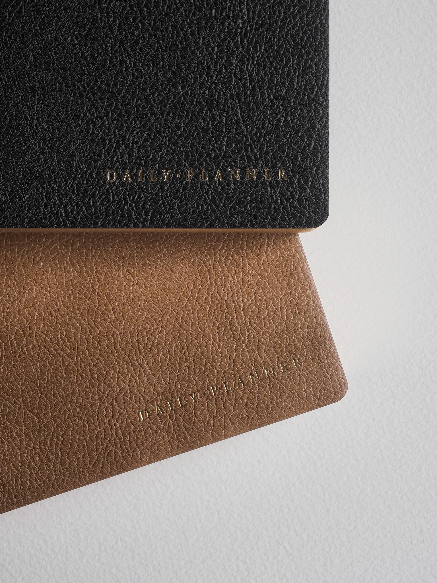 Two textured daily planners in contrasting black and natural tan faux leather, both featuring the embossed "DAILY PLANNER" title.
