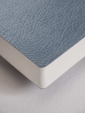 Close-up of a notebook or book corner showing a textured dusty blue cover and white page edges on a light gray surface.
#Color_Mist