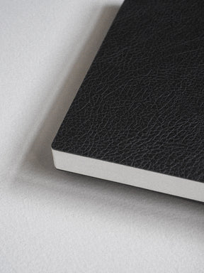 A close-up of a black textured notebook or journal corner with white page edges, resting on a light gray surface.
#Color_Onyx