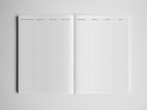 Open academic planner featuring white gridded pages with structured layouts for goal breakdowns, quarterly, monthly, weekly, and daily planning.