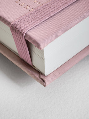 A close-up of a hardcover notebook with a dusty pink fabric cover and matching elastic closure, showing white pages and a textured surface.
#Color_Seashell