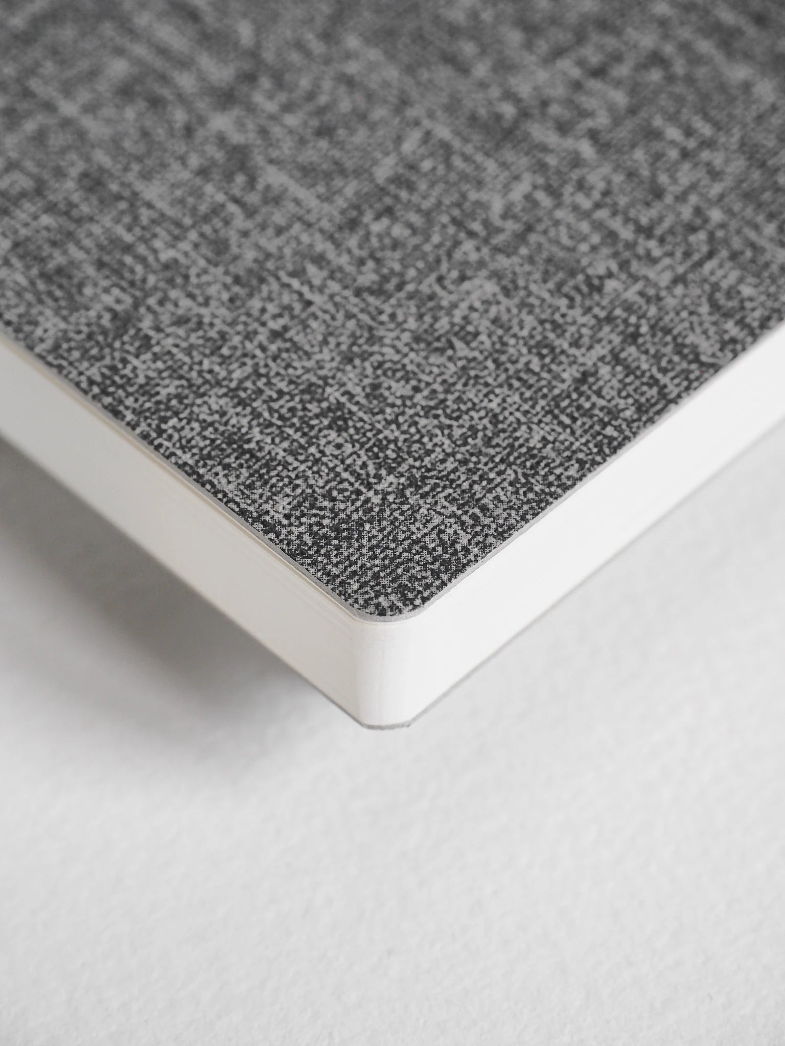 A close-up of a notebook or book corner showing a textured gray cover and white page edges on a light neutral background.
#Color_Tweed