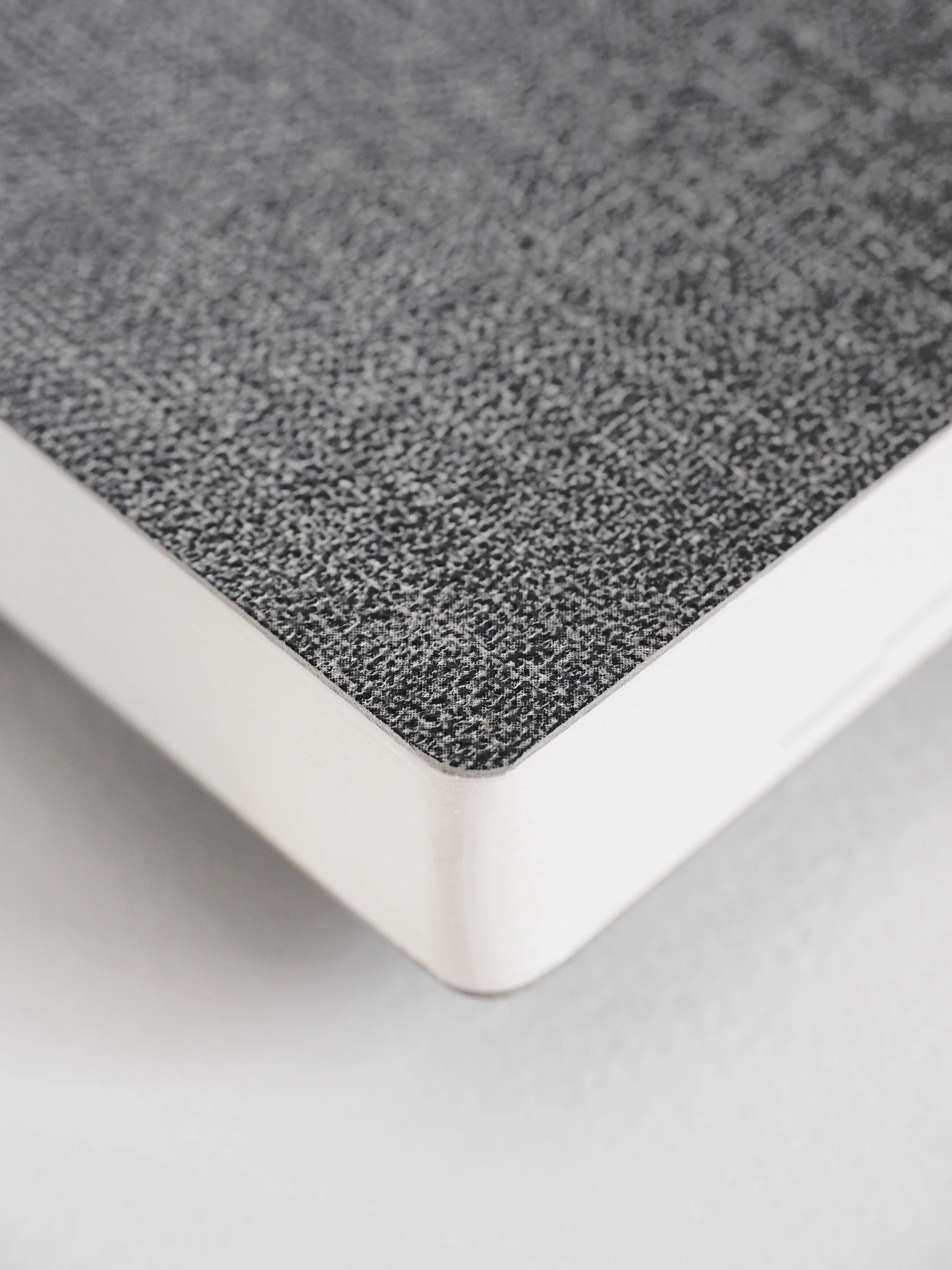 A close-up of a notebook or book corner showing a textured gray cover and white page edges on a light neutral background.
#Color_Tweed