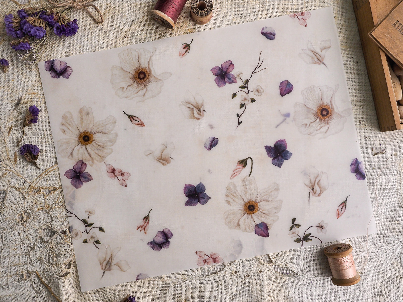 Pale vellum paper featuring a delicate, scattered botanical pattern of cream and muted purple dried flowers and subtle floral sketches.
