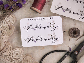 Elegant white sticker displaying "February" in cursive script, adorned with delicate watercolor florals, small moths, and the "Sterling Ink" brand text.