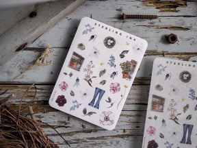 Pastel spring-themed sticker collection featuring delicate floral icons, potted greenery, a bird's nest, and miniature denim jeans.
