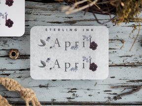 Elegant botanical sticker for April 2023 featuring the word "April" in serif text and delicate decorative accents of purple flowers and sepia-toned leaves.