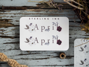 Delicate botanical sticker featuring the word April alongside clusters of soft purple and white watercolor flowers.