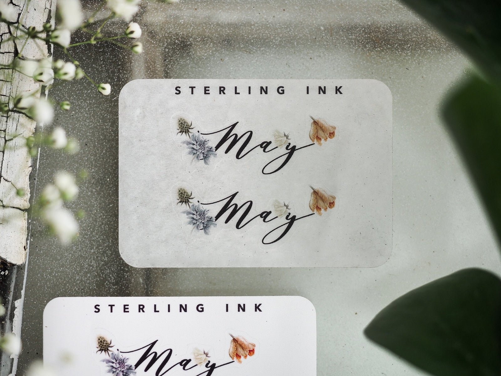 May month sticker featuring scripted lettering, scattered purple florals, and dried beige botanical accents for seasonal planning and journaling.