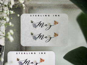 May month sticker featuring scripted lettering, scattered purple florals, and dried beige botanical accents for seasonal planning and journaling.