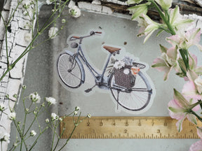 Vintage die-cut sticker featuring an artistic, monochromatic bicycle carrying a rustic basket filled with detailed spring blooms.